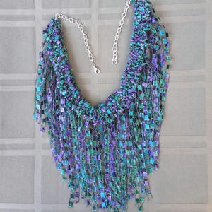 K&ouml;nnte beinhalten: Eine handgefertigte Halskette mit einem kaskadierenden Design. Die Halskette hat ein gewebtes, strukturiertes L&auml;tzchen in T&uuml;rkis-, Lila- und Gr&uuml;nt&ouml;nen mit herabh&auml;ngenden Str&auml;ngen aus quadratischen Perlen. Die Halskette hat eine silberne Kette und einen Verschluss.