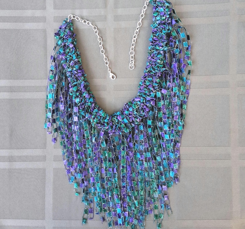 K&ouml;nnte beinhalten: Eine handgefertigte Halskette mit einem kaskadierenden Design. Die Halskette hat ein gewebtes, strukturiertes L&auml;tzchen in T&uuml;rkis-, Lila- und Gr&uuml;nt&ouml;nen mit herabh&auml;ngenden Str&auml;ngen aus quadratischen Perlen. Die Halskette hat eine silberne Kette und einen Verschluss.