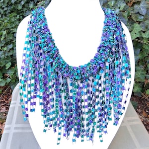 K&ouml;nnte beinhalten: Eine handgefertigte Statement-Halskette mit gewebtem Design. Die Halskette hat eine lebendige Farbpalette aus T&uuml;rkis, Lila und Gr&uuml;n. Die fransenartigen Str&auml;nge h&auml;ngen von einem strukturierten Kragen herab und schaffen ein einzigartiges Accessoire.