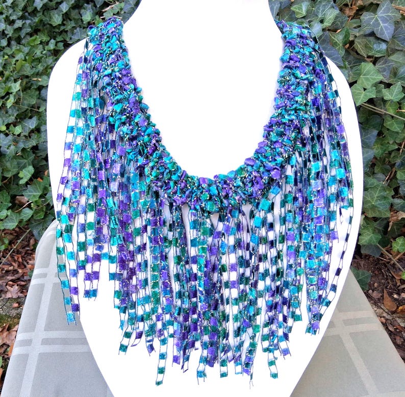 K&ouml;nnte beinhalten: Eine handgefertigte Statement-Halskette mit gewebtem Design. Die Halskette hat eine lebendige Farbpalette aus T&uuml;rkis, Lila und Gr&uuml;n. Die fransenartigen Str&auml;nge h&auml;ngen von einem strukturierten Kragen herab und schaffen ein einzigartiges Accessoire.