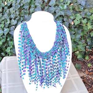 K&ouml;nnte beinhalten: Eine handgefertigte Halskette mit Fransen-Design. Die Halskette hat einen gewebten Kragen in Lila-, T&uuml;rkis- und Blaut&ouml;nen mit langen, baumelnden Fransen in passenden Farben. Die Halskette wird auf einer wei&szlig;en Schaufensterpuppe pr&auml;sentiert.