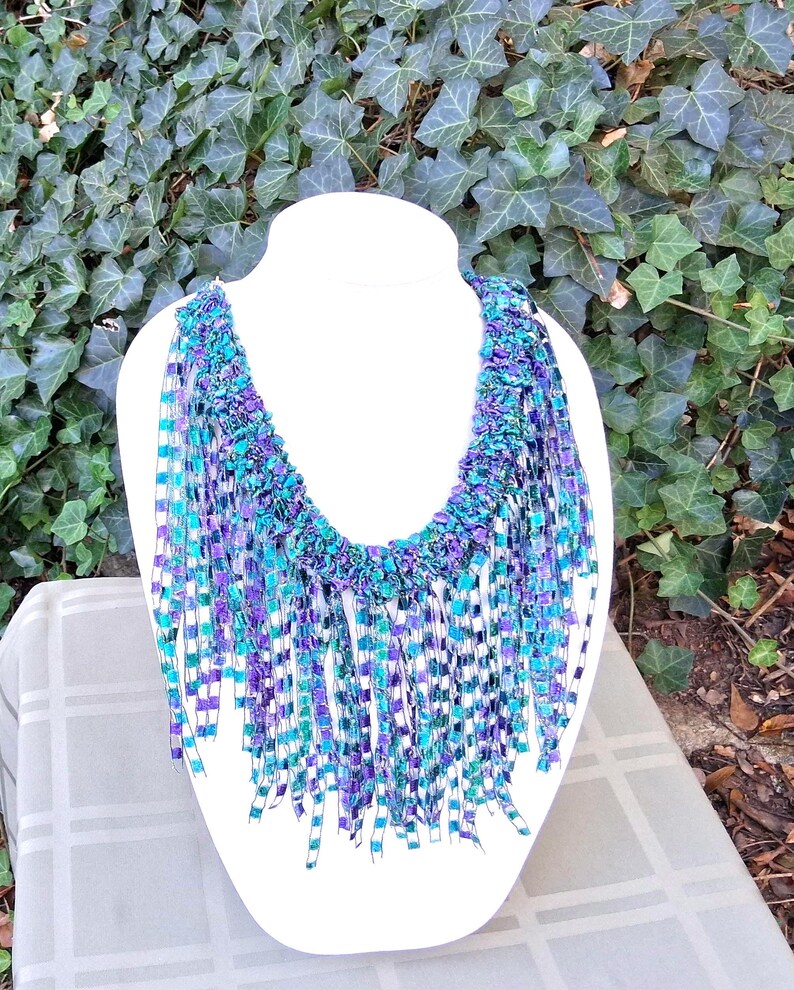 K&ouml;nnte beinhalten: Eine handgefertigte Halskette mit Fransen-Design. Die Halskette hat einen gewebten Kragen in Lila-, T&uuml;rkis- und Blaut&ouml;nen mit langen, baumelnden Fransen in passenden Farben. Die Halskette wird auf einer wei&szlig;en Schaufensterpuppe pr&auml;sentiert.