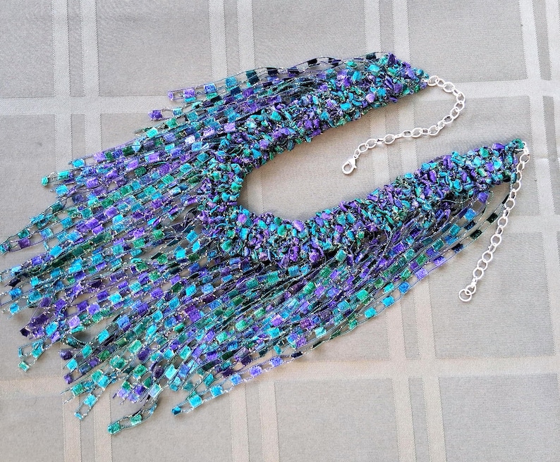 K&ouml;nnte beinhalten: Handgefertigte Halskette mit einzigartigem Design. Die Halskette hat eine lebendige Farbmischung, darunter Blau-, Lila- und Gr&uuml;nt&ouml;ne. Sie hat ein strukturiertes, fransiges Aussehen und wird durch eine silberne Kette akzentuiert.