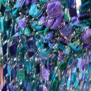 K&ouml;nnte beinhalten: Nahaufnahme eines Textils mit einer Mischung aus Texturen und Farben. Das St&uuml;ck weist eine Kombination aus t&uuml;rkis-, lila- und blaufarbenen F&auml;den mit einem fransenartigen Effekt auf. Das Design umfasst strukturierte, kn&ouml;tchenartige Elemente und schimmernde Akzente.