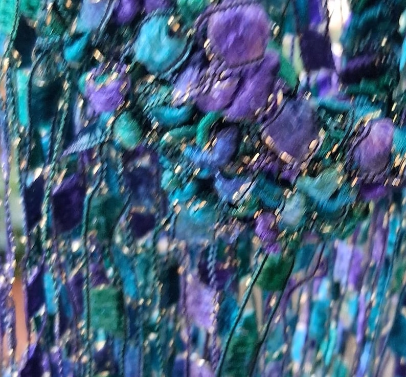 K&ouml;nnte beinhalten: Nahaufnahme eines Textils mit einer Mischung aus Texturen und Farben. Das St&uuml;ck weist eine Kombination aus t&uuml;rkis-, lila- und blaufarbenen F&auml;den mit einem fransenartigen Effekt auf. Das Design umfasst strukturierte, kn&ouml;tchenartige Elemente und schimmernde Akzente.