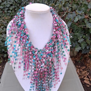 May include: A colorful, handmade necklace displayed on a white mannequin bust. The necklace features a mix of pink, blue, and green fabric strips, creating a textured and vibrant accessory. The fringe-like design adds a unique touch to this statement piece.