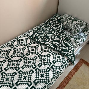 May include: A bed with a white frame is covered with a green and white patterned blanket and pillow. The blanket features a geometric design. A patterned pillow with a green and white design rests on the bed. A rug with a brown border is visible.