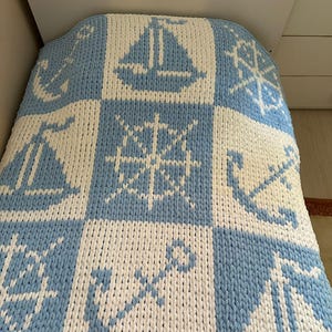 Op de afbeelding: Een dikke gebreide deken met een nautisch thema. De deken heeft een patroon van blauwe en witte vierkanten, elk met een zeilboot, scheepsrad of anker. De deken is gemaakt van een zacht, dik garen.