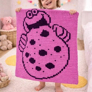 May include: A knitted pink blanket featuring a cookie design with dark purple accents. The blanket is held up by a child in a room with a white rug and a pink teddy bear.