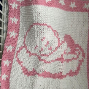 May include: A handmade baby blanket in pink and white. The blanket features a pink border with white star accents. The center of the blanket has a pink outline of a baby. The blanket is made of a soft, chunky knit.