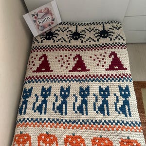 以下が含まれることがあります： クリーム色の、ハロウィーンをテーマにしたニットブランケット。黒いクモ、魔女の帽子、青い猫、オレンジ色のジャック・オー・ランタンが特徴です。ブランケットは繰り返し模様で、太い糸でできています。「Knitly」というブランド名が確認できます。