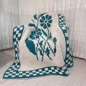 Puede incluir: Una manta de punto blanca y azul verdoso con un borde a cuadros y un diseño floral. La manta está drapeada sobre una silla.