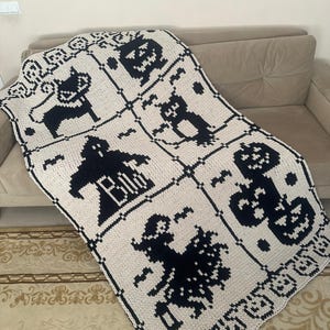 May include: A cream-colored Halloween-themed knitted blanket with black graphic designs. The blanket features a grid pattern with images of a black cat, a ghost with the word "Boo", a jack-o'-lantern, and a witch. The blanket is draped over a beige sofa.