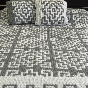 May include: A gray and white knitted bedspread with two matching square pillows. The pillows and bedspread feature a geometric pattern. The color scheme is a neutral gray and white, creating a modern and cozy aesthetic.