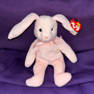 Può includere: Coniglio di peluche rosa con orecchie lunghe e un'etichetta Ty. Il coniglio è seduto su una superficie viola.