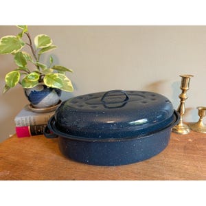 May include: An oval-shaped dark blue roasting pan with a speckled white enamel finish and a matching lid. The roaster features two side handles and a central handle on the lid. The exterior surface has a glossy, mottled appearance throughout.