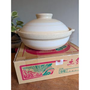 May include: A beige and white ceramic donabe pot with a matching lid, featuring horizontal stripes and two side handles. The pot sits on a cardboard box decorated with colorful illustrations of food and Japanese text, including the label 9号.