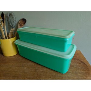 May include: Two rectangular, mint green plastic storage containers with matching lids stacked on top of each other. The containers have a smooth, solid finish and a simple, functional design, positioned next to a yellow utensil holder with kitchen tools.