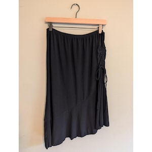 May include: A solid black skirt featuring an elasticized waistband and a side drawstring detail that creates a gathered effect. The skirt has an asymmetrical hemline with a diagonal seam across the lower portion, creating a flared, flowing silhouette.