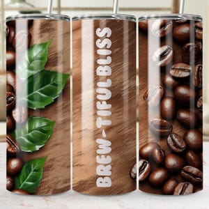 Puede incluir: Tres vasos de acero inoxidable con diferentes diseños. El primer vaso presenta un fondo de madera con hojas verdes y granos de café. El segundo vaso tiene un fondo de madera con el texto "Brew-tiful Bliss". El tercer vaso tiene un fondo de madera con granos de café.