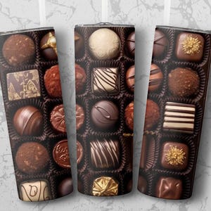 Pode incluir: Três canecas isoladas com um padrão repetido de bombons sortidos numa caixa. Os bombons são de várias formas e cores, incluindo chocolate preto e de leite, alguns decorados com detalhes dourados.