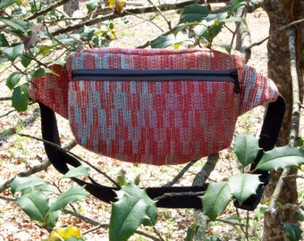 May include: A red and blue patterned fanny pack with a black zip and adjustable strap. The bag is made of woven fabric and is shown hanging in a tree. The fanny pack is a hands-free bag for carrying essentials.