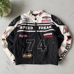 May include: Black and cream-colored racing jacket with multiple sponsor patches and the words "SPEED FREAK" and "74" prominently displayed. The jacket features a stand-up collar, a zipper closure, and ribbed cuffs and hem. The sleeves have the words "RACE TRACK".