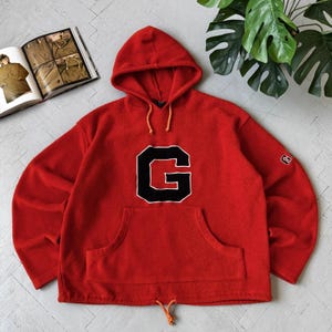 Peut inclure: Un sweat à capuche en polaire rouge vif arborant une grande lettre G noire avec un contour blanc centrée sur la poitrine. Le vêtement comprend une poche kangourou, une capuche à cordon de serrage orange et un petit logo G sur la manche gauche.