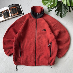 May include: A red fleece jacket with a black collar and zipper closure. The jacket features two side pockets with black zippers and the brand logo on the chest. The jacket is made of a soft, textured material.