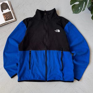 May include: A black and blue fleece jacket with a full zipper closure. The jacket has a high collar and two zippered pockets. The North Face logo is on the upper left chest. The sleeves and lower portion of the jacket are blue.