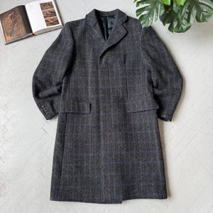 Op de afbeelding: Een lange, donkergrijze tweed jas met een subtiel blauw ruitpatroon. De jas heeft een reverskraag, twee voorzakken en manchetten met knopen. De jas ligt plat op een lichtgekleurd oppervlak en toont zijn op maat gemaakte ontwerp.