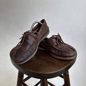 May include: A pair of dark brown leather deck shoes featuring a moccasin-style stitched toe, metal eyelets, and thin leather laces. The shoes have a low-profile silhouette with a flat rubber sole and a visible metal shoe tree inside the left shoe.