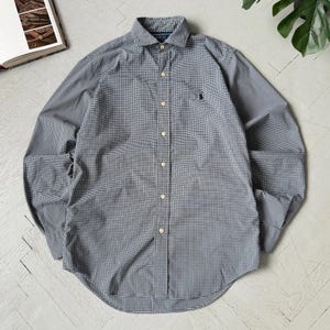 May include: A long-sleeved button-down shirt in a blue and white checkered pattern. The shirt features a classic collar, a chest pocket, and a small embroidered logo. The shirt is made of a lightweight fabric.