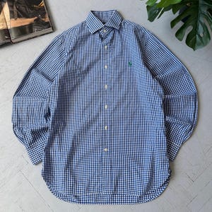May include: A long-sleeved button-down shirt in a blue and white gingham pattern. The shirt has a classic collar and a small green logo on the chest. The shirt is made of a lightweight fabric.