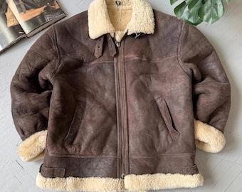 May include: A dark brown leather jacket featuring a thick cream-colored shearling lining, a wide fold-over collar, a full-length front zipper, two side pockets, and adjustable buckle straps at the collar and hem for a secure and structured fit.