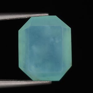 May include: A rectangular, emerald-cut gemstone with a smooth, translucent surface in a soft seafoam green color. The stone features beveled edges and a flat top facet, held securely by the metal tips of a pair of tweezers against a black background.