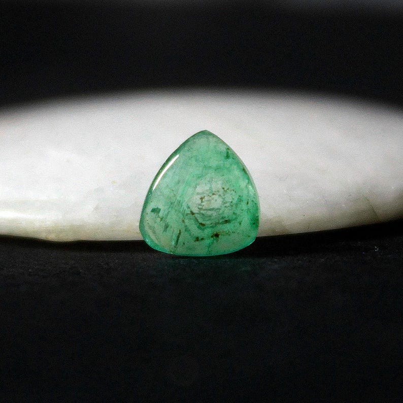 May include: A triangular, polished emerald gemstone with a translucent, vibrant green hue. The gem displays internal patterns and inclusions, resting on a white, rounded surface against a dark background. The gemstone is a cabochon cut.