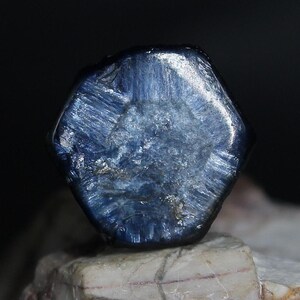 May include: A close-up of a faceted, hexagonal, deep blue gemstone. The gem has a textured surface with visible striations and a slightly lighter, almost white, interior. The stone is set against a dark background and a light-colored, textured surface.