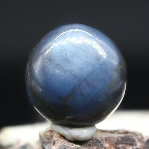 May include: A polished, spherical gemstone with a deep blue hue. The gem has a smooth, reflective surface and sits atop a small, light-colored base. The stone's color transitions from dark blue to lighter shades, creating a subtle gradient.