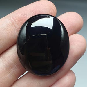 May include: A polished, oval-shaped black gemstone with a glossy surface. The stone reflects light, creating bright highlights. The smooth, dark surface suggests a cabochon or a similar type of polished stone, suitable for jewelry or decorative purposes.