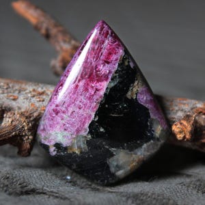 May include: A faceted gemstone with a triangular shape. The stone features a striking combination of colors, including deep magenta, black, and hints of beige. The magenta section has a textured appearance, while the black area is smooth.