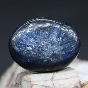 May include: A polished, oval-shaped gemstone with a deep blue hue and a shimmering, fibrous internal structure. The stone is set against a neutral background, highlighting its natural beauty and intricate patterns. The gemstone is a unique piece of jewelry.