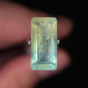 May include: A rectangular, emerald-cut gemstone with a pale yellow-green hue and visible internal inclusions. The stone is set in a simple metal ring band, showcasing a translucent quality and faceted edges that catch the light against a dark background.