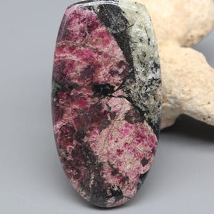 May include: A polished, oval-shaped gemstone cabochon featuring a vibrant mix of deep magenta, dark black, and translucent white mineral inclusions. The surface displays a smooth, glossy finish with distinct natural patterns and crystalline textures.