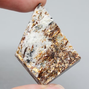 May include: A rhomboid-shaped gemstone with a mix of white, brown, and gold colors. The stone has a textured surface with visible crystalline structures. The gemstone is held between two fingers against a gray background.