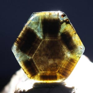 May include: A hexagonal gemstone with a translucent, layered appearance. The gem features a mix of brown, yellow, and blue-green hues, with darker brown hexagonal patterns within. The stone is well-lit, showcasing its internal structure and clarity.
