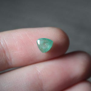 May include: A small, triangular, polished emerald gemstone. The gem is a vibrant green color and appears translucent. The stone is held between two fingers, with a neutral background.
