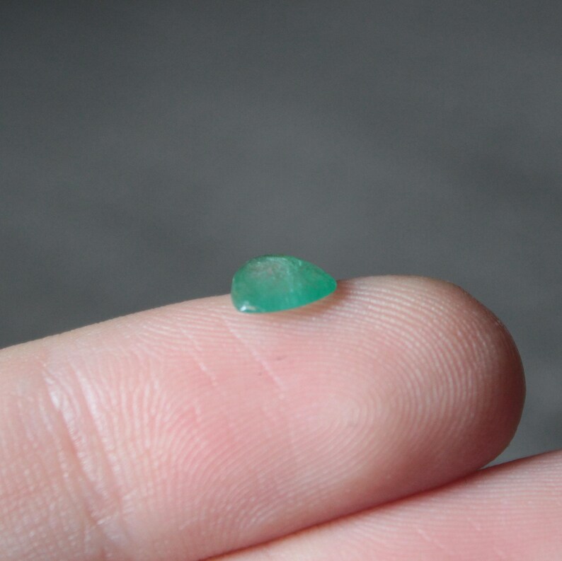 May include: A small, teardrop-shaped, emerald-green gemstone. The gem is set on a fingertip, showcasing its size and color against the skin. The background is a muted gray, providing contrast to the vibrant jewel.