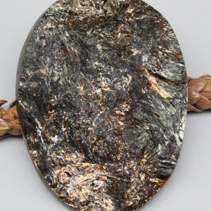 May include: Oval-shaped gemstone pendant with a textured surface. The stone features a mix of dark gray, black, and gold colors, creating a unique pattern. The pendant is likely for jewelry making or as a decorative element.