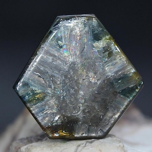 May include: A faceted, iridescent gemstone with a hexagonal shape. The stone displays a range of colors, including silver, blue, and green, with internal striations. The gemstone is set against a dark background.